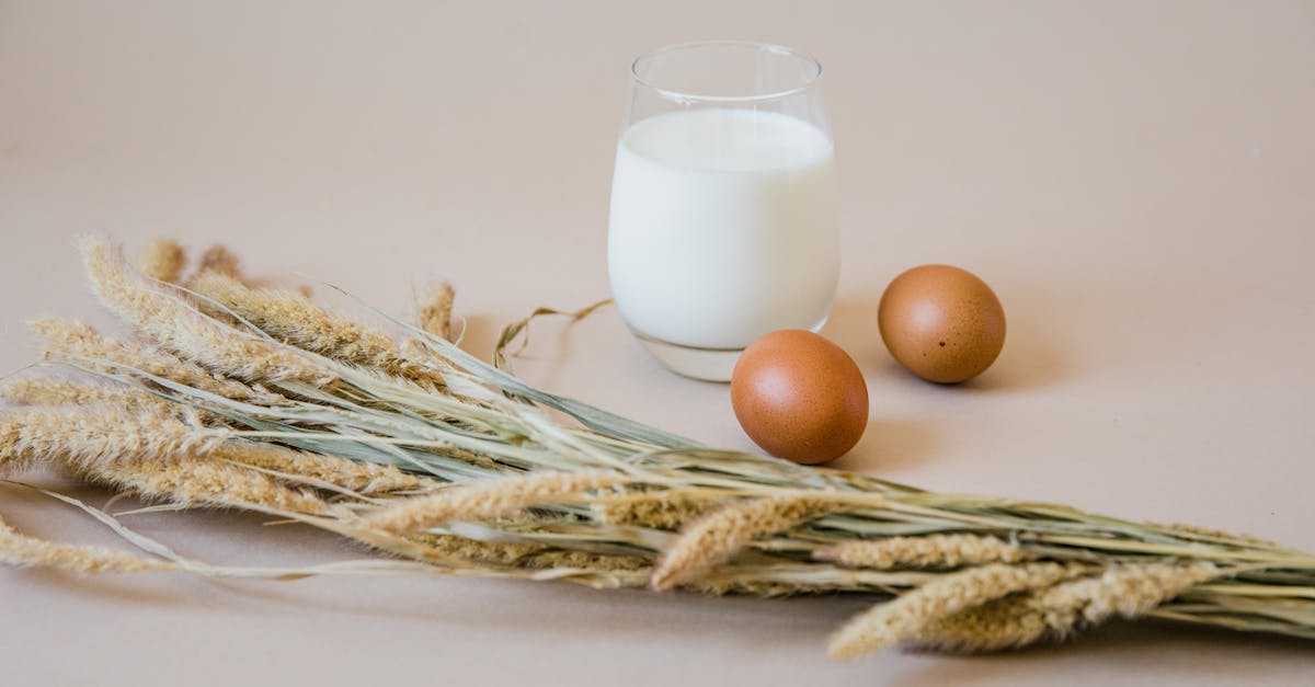 Still life of a glass of milk, brown eggs, and wheat stalks on a beige background.